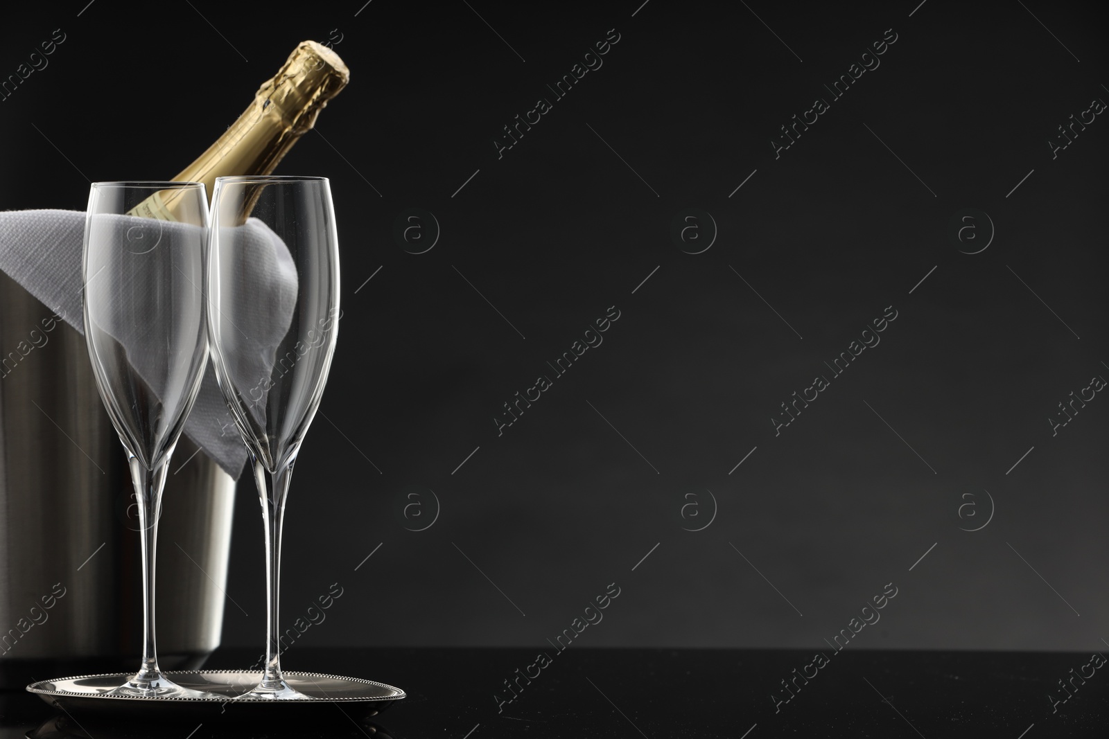 Photo of Bottle of sparkling wine in cooler bucket and glasses on black table against grey background. Space for text