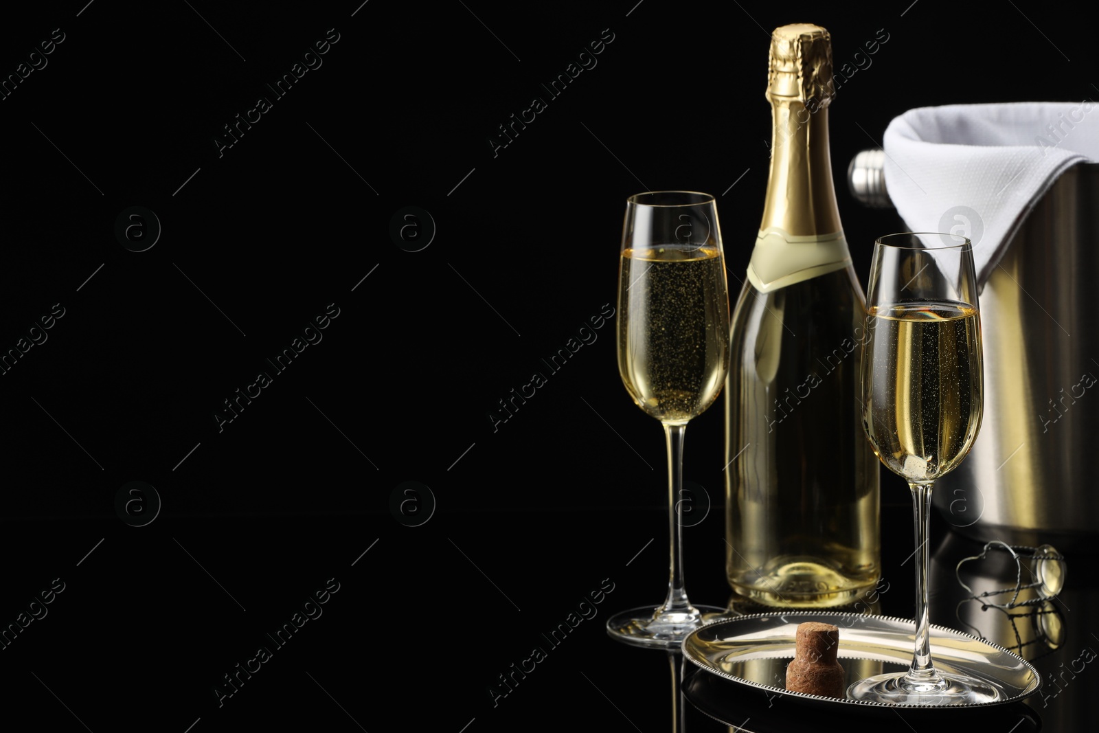 Photo of Bottle of sparkling wine, glasses and cooler bucket on black mirror surface. Space for text