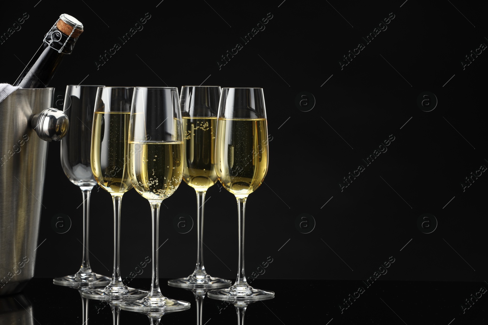 Photo of Bottle of sparkling wine in cooler bucket and glasses on black mirror surface. Space for text