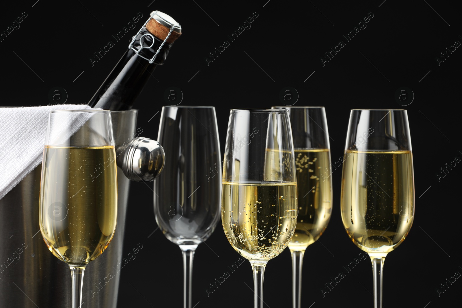 Photo of Bottle of sparkling wine in cooler bucket and glasses against black background, closeup