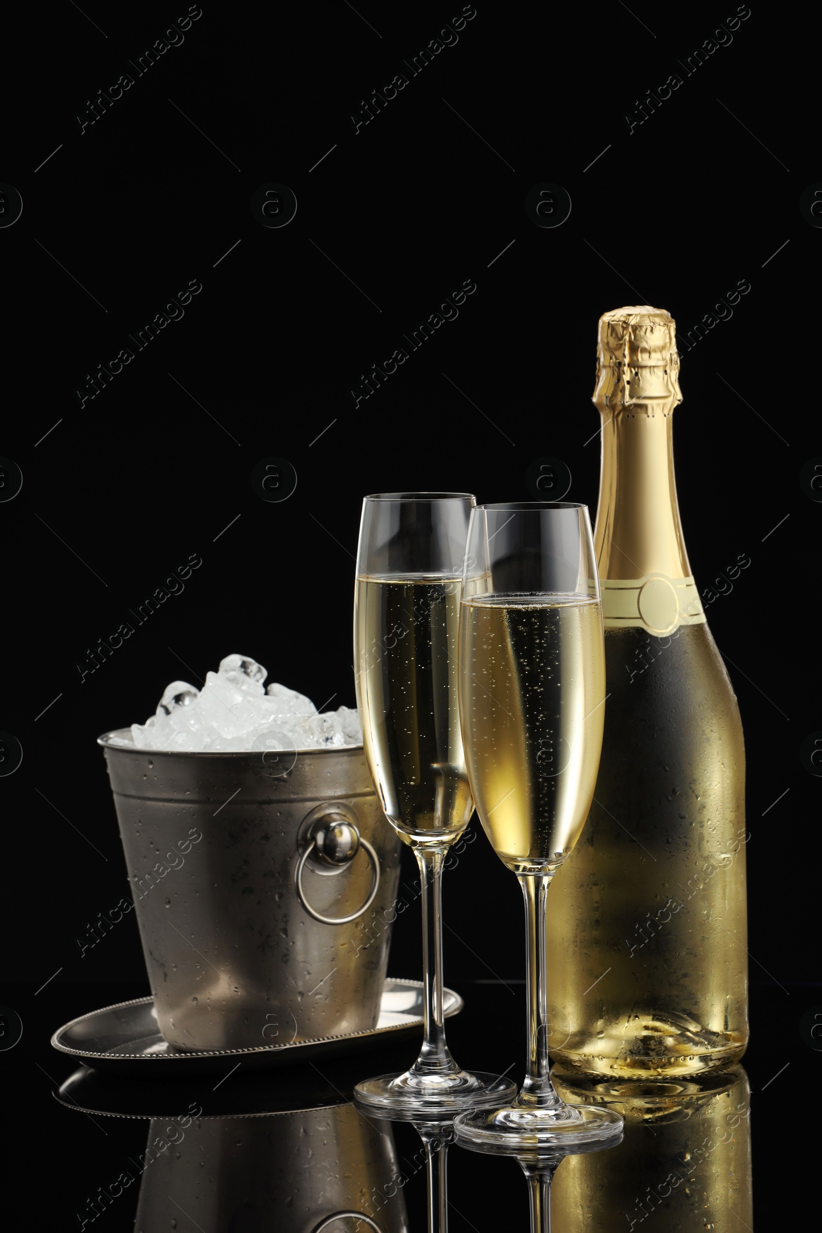 Photo of Bottle of sparkling wine, glasses and cooler bucket on black mirror surface