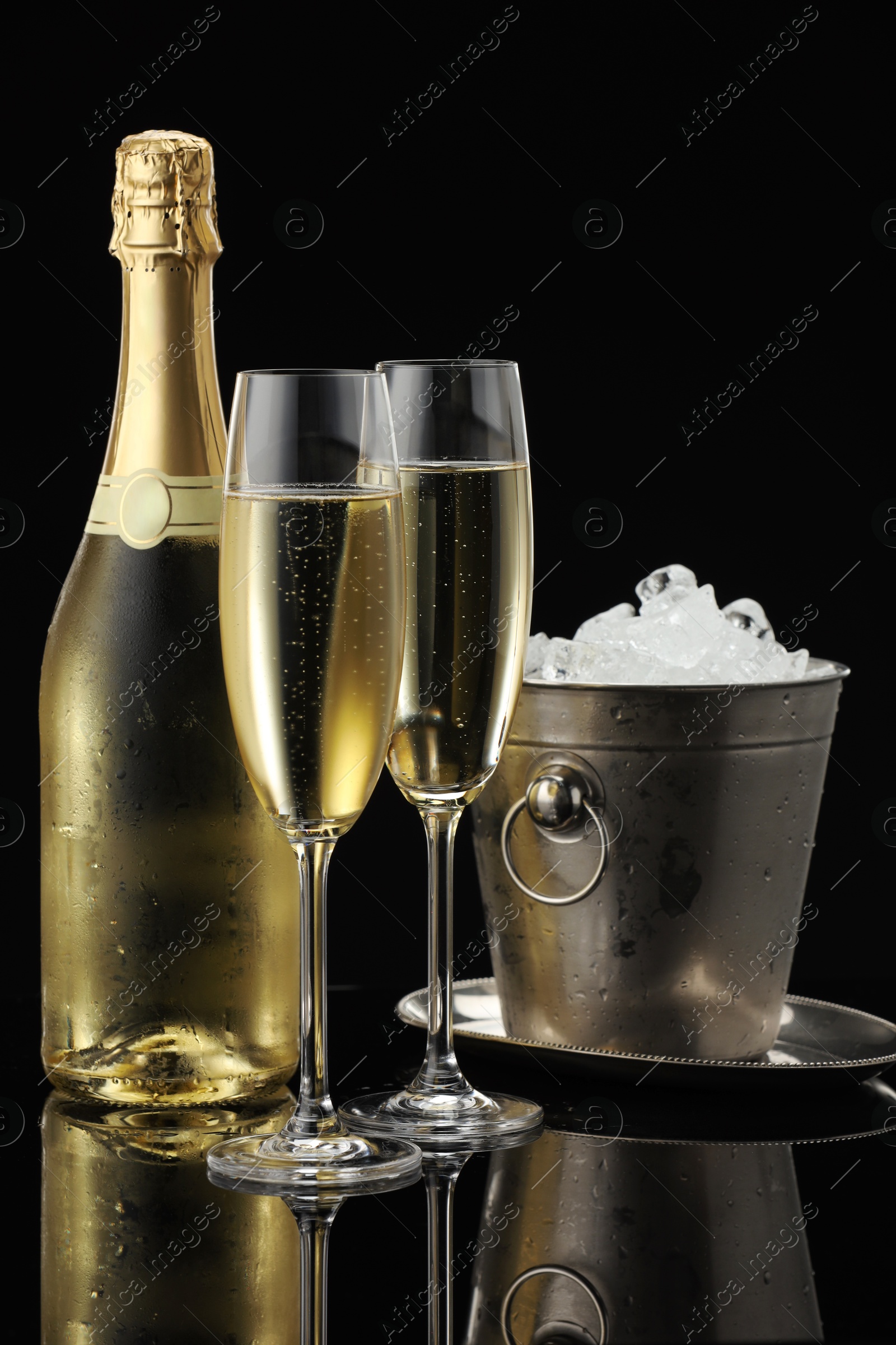 Photo of Bottle of sparkling wine, glasses and cooler bucket on black mirror surface