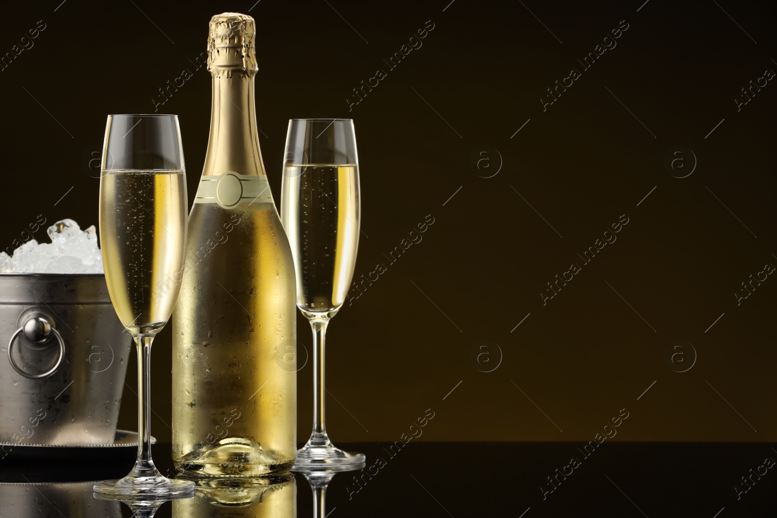 Photo of Bottle of sparkling wine, glasses and cooler bucket on black mirror surface. Space for text