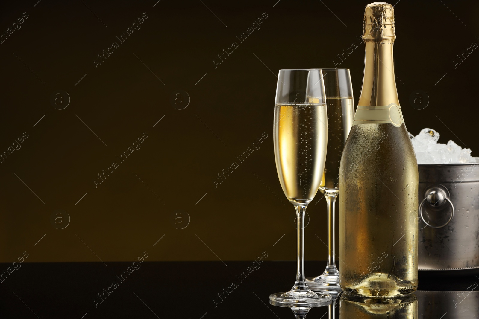 Photo of Bottle of sparkling wine, glasses and cooler bucket on black mirror surface. Space for text