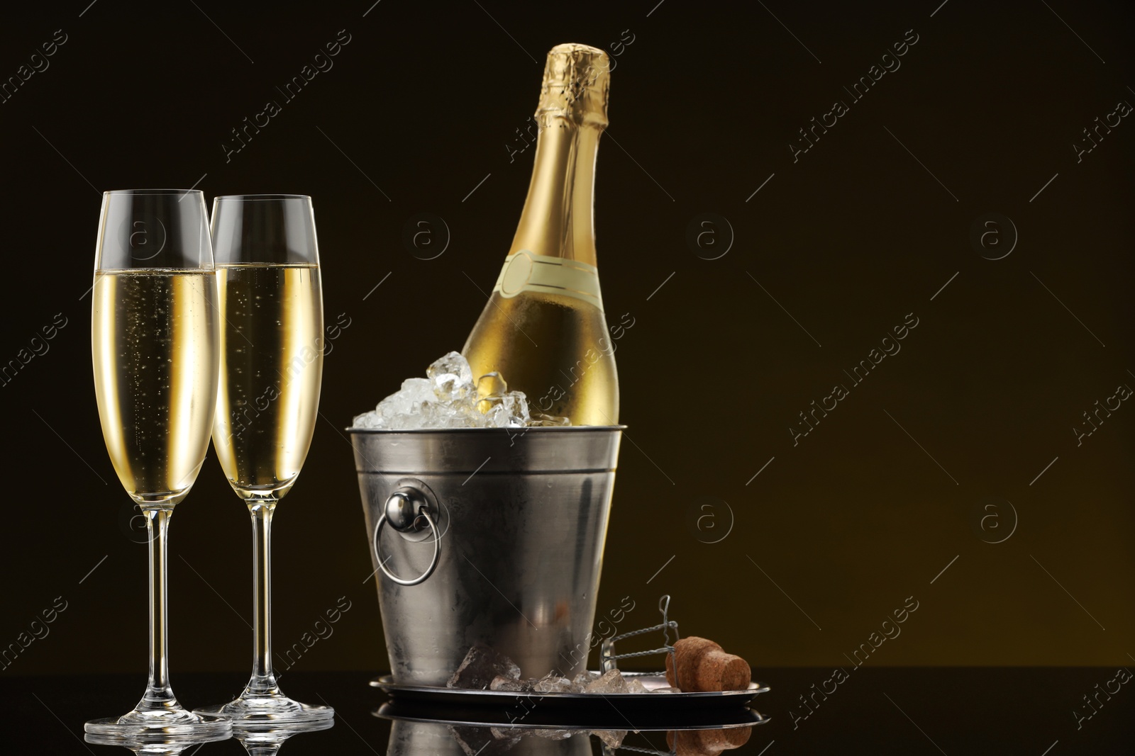 Photo of Bottle of sparkling wine in cooler bucket and glasses on black mirror surface. Space for text