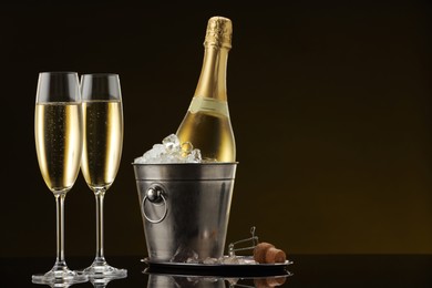 Photo of Bottle of sparkling wine in cooler bucket and glasses on black mirror surface. Space for text