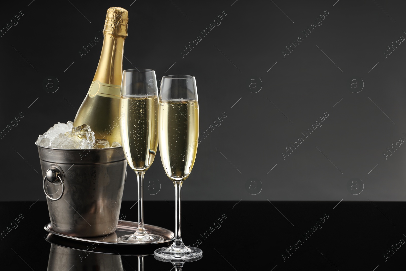 Photo of Bottle of sparkling wine in cooler bucket and glasses on black mirror surface. Space for text