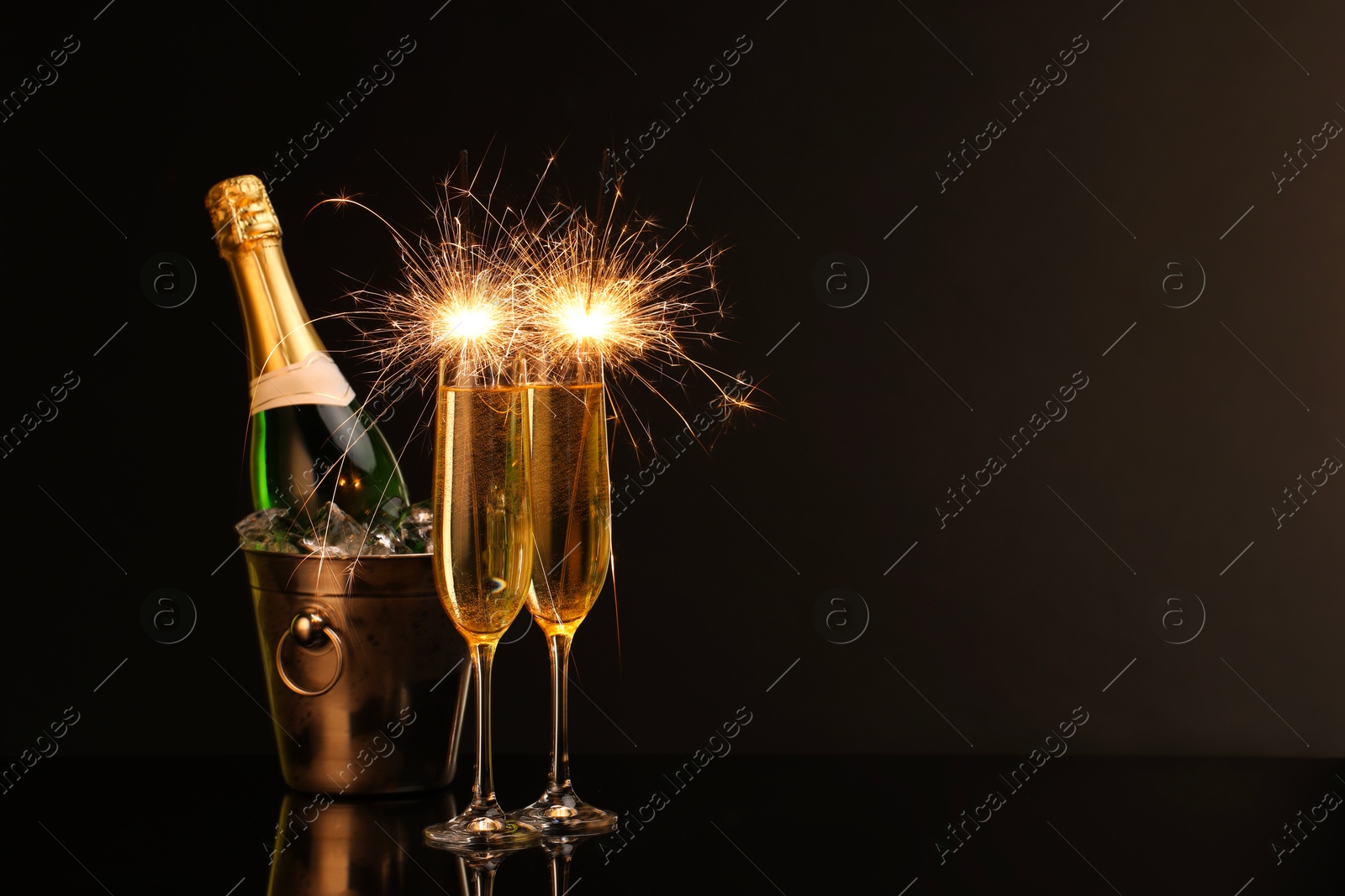 Photo of Glasses of sparkling wine with sparklers and bottle in cooler bucket on black background. Space for text