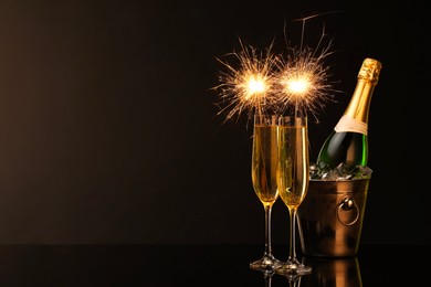 Photo of Glasses of sparkling wine with sparklers and bottle in cooler bucket on black background. Space for text