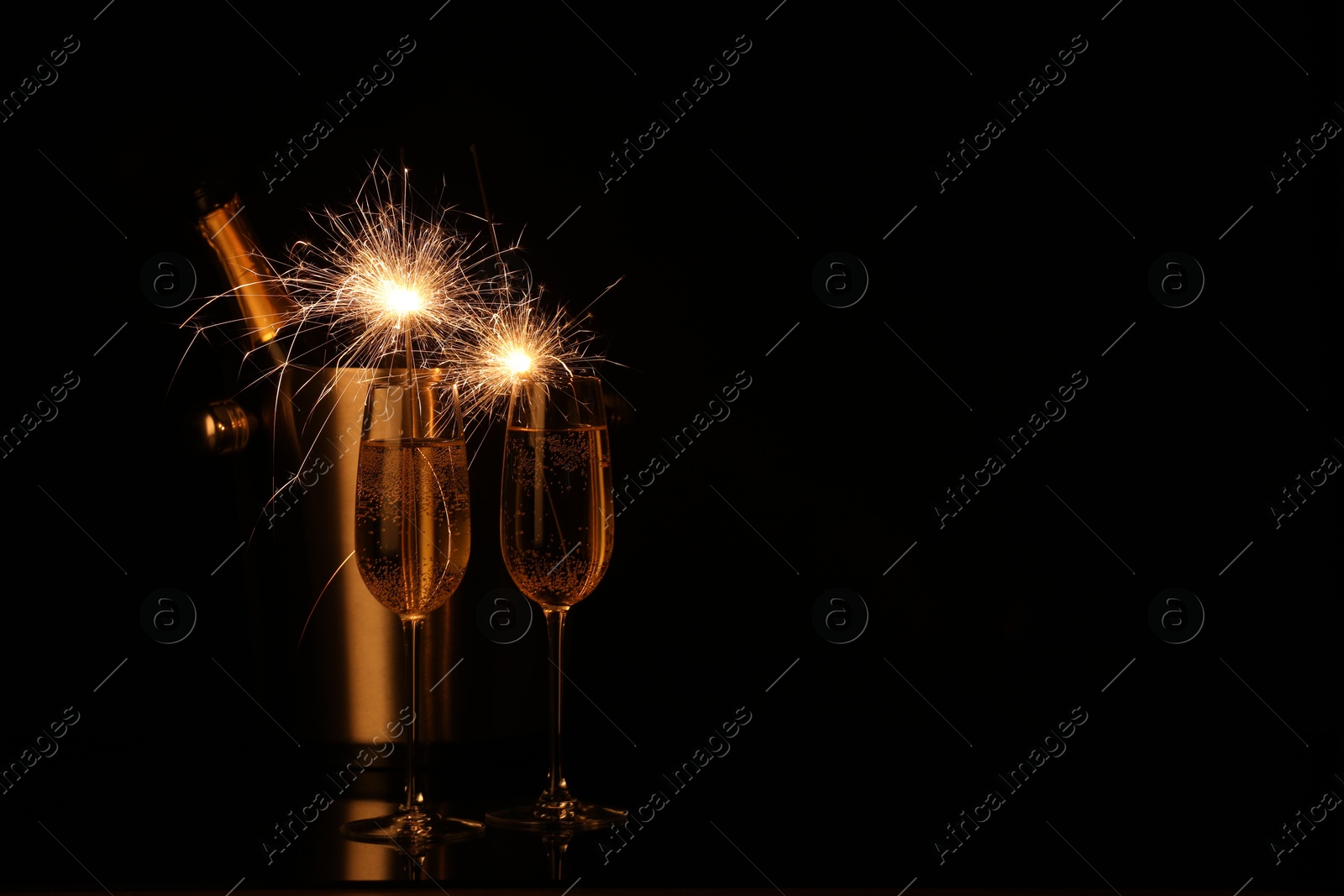 Photo of Glasses of sparkling wine with sparklers and bottle in cooler bucket on black background. Space for text