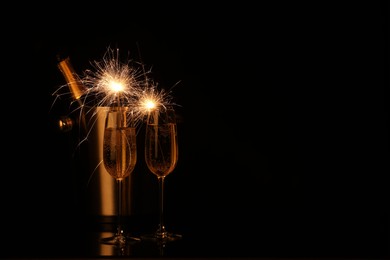 Photo of Glasses of sparkling wine with sparklers and bottle in cooler bucket on black background. Space for text