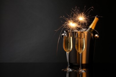 Glasses of sparkling wine with sparklers and bottle in cooler bucket on black background. Space for text