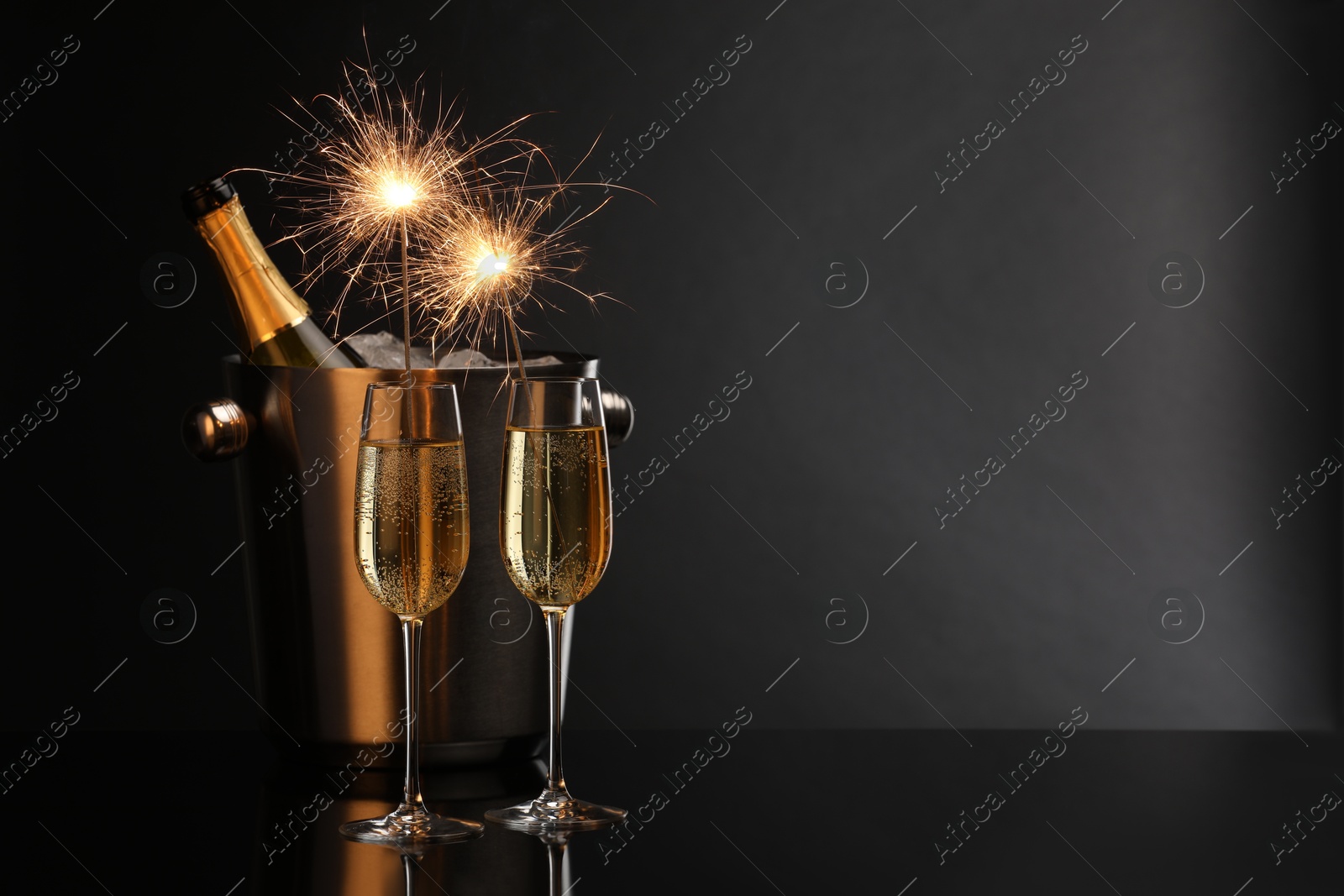 Photo of Glasses of sparkling wine with sparklers and bottle in cooler bucket on black background. Space for text