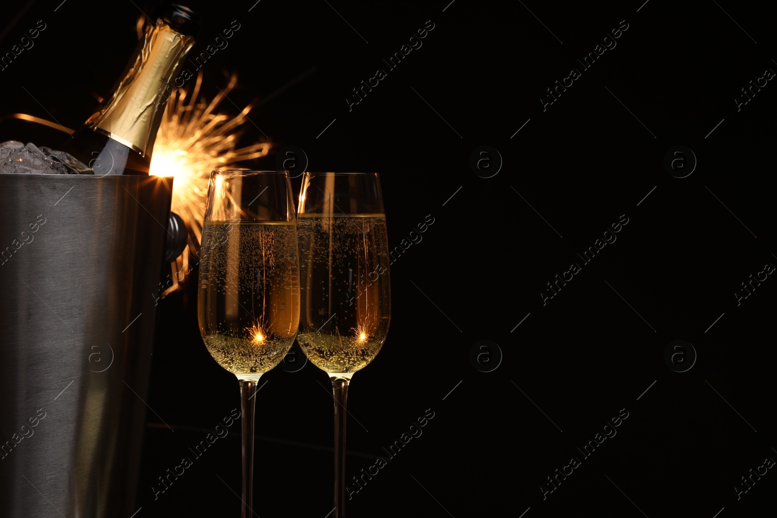 Photo of Glasses of sparkling wine with sparklers and bottle in cooler bucket on black background, closeup. Space for text