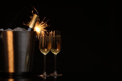 Photo of Glasses of sparkling wine with sparklers and bottle in cooler bucket on black background. Space for text