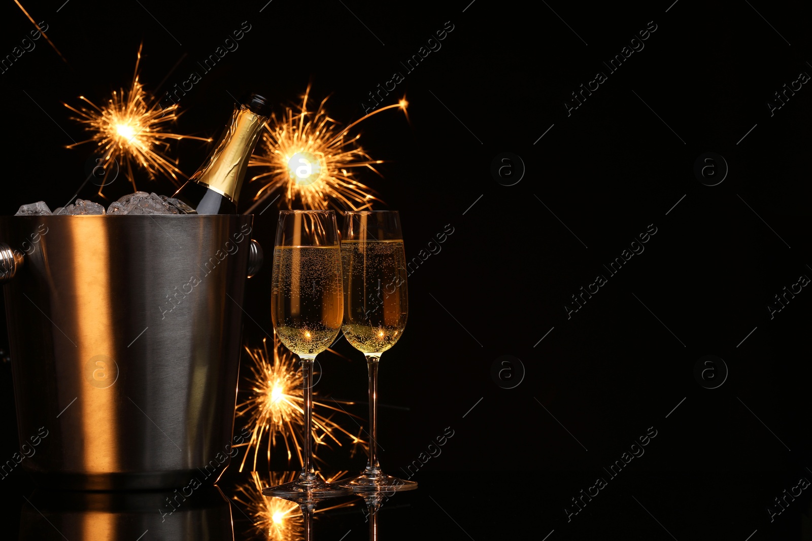 Photo of Glasses of sparkling wine with sparklers and bottle in cooler bucket on black background. Space for text