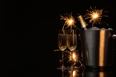 Photo of Glasses of sparkling wine with sparklers and bottle in cooler bucket on black background. Space for text