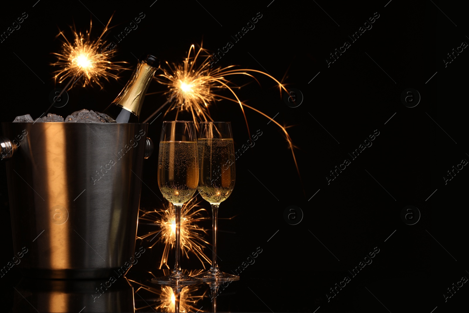 Photo of Glasses of sparkling wine with sparklers and bottle in cooler bucket on black background. Space for text