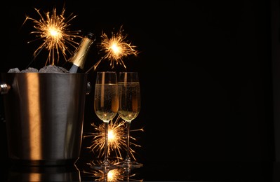 Photo of Glasses of sparkling wine with sparklers and bottle in cooler bucket on black background. Space for text