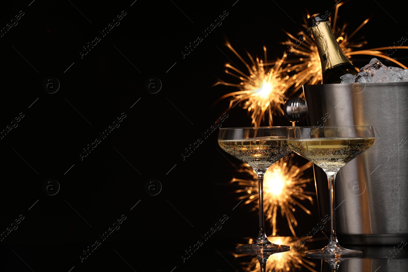 Photo of Glasses of sparkling wine with sparklers and bottle in cooler bucket on black background. Space for text