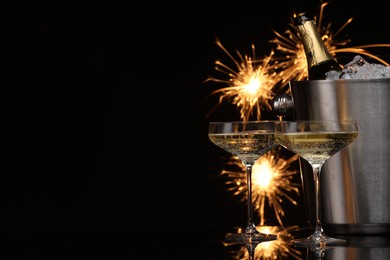 Photo of Glasses of sparkling wine with sparklers and bottle in cooler bucket on black background. Space for text