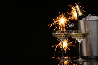 Photo of Glasses of sparkling wine with sparklers and bottle in cooler bucket on black background. Space for text