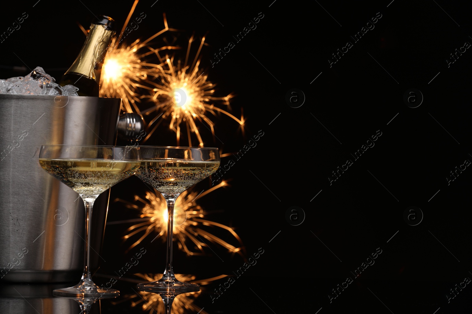 Photo of Glasses of sparkling wine with sparklers and bottle in cooler bucket on black background. Space for text