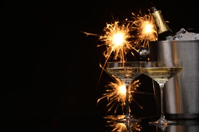 Photo of Glasses of sparkling wine with sparklers and bottle in cooler bucket on black background. Space for text