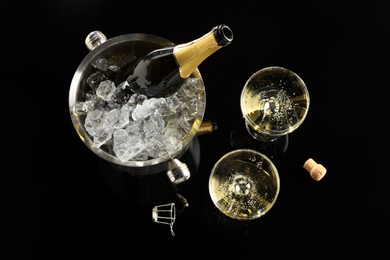 Photo of Bottle of sparkling wine in cooler bucket and glasses on black table, flat lay