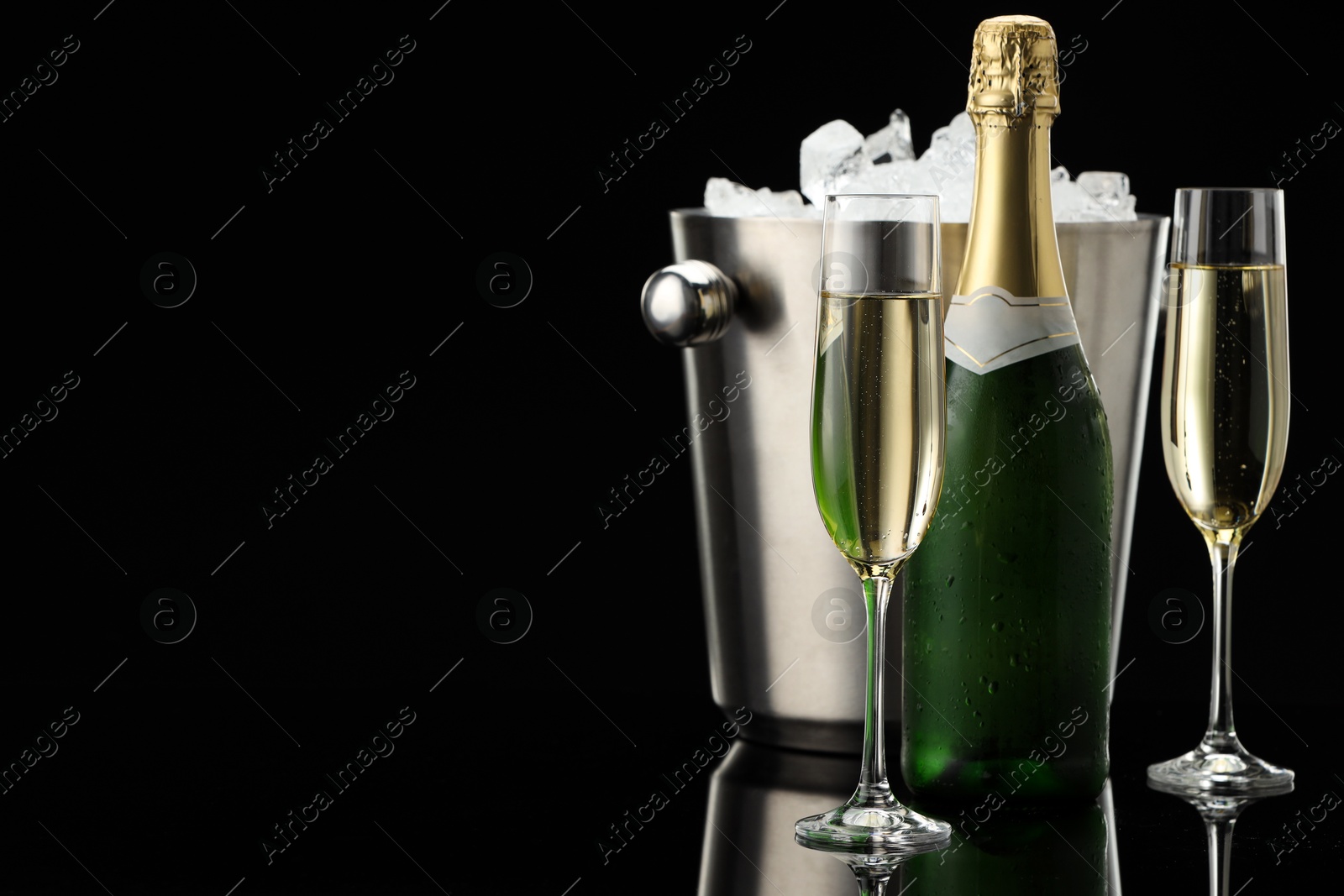 Photo of Bottle of sparkling wine, glasses and cooler bucket on black mirror surface. Space for text