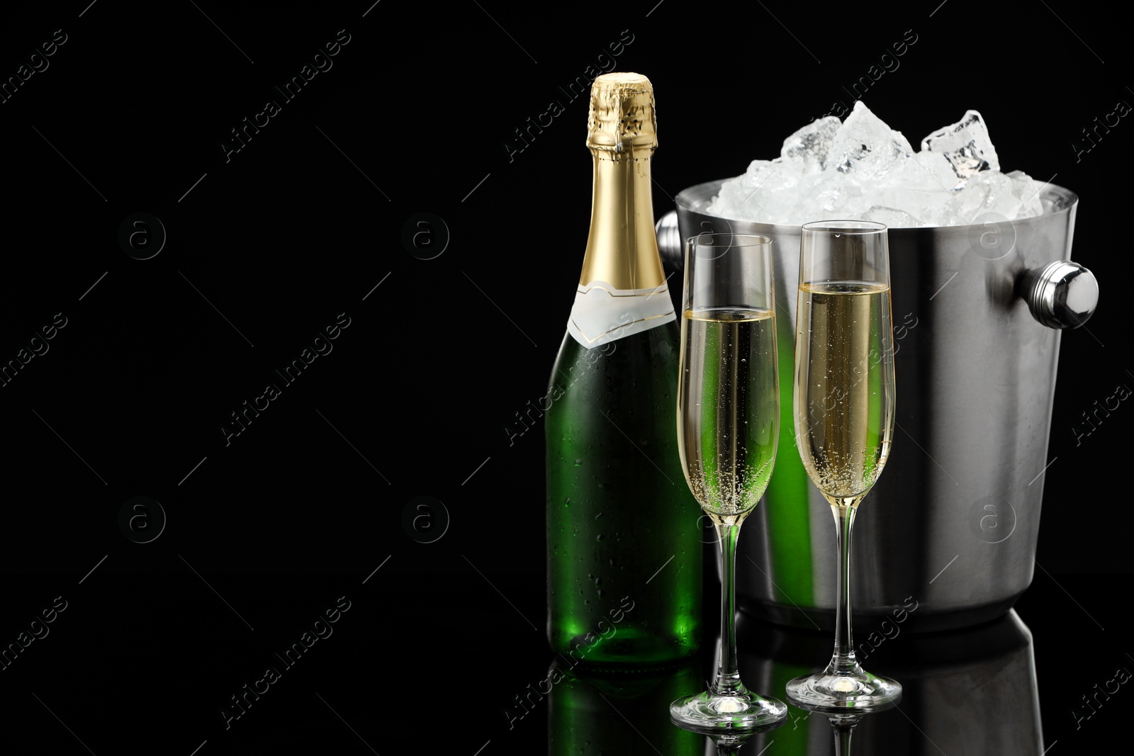 Photo of Bottle of sparkling wine, glasses and cooler bucket on black mirror surface. Space for text