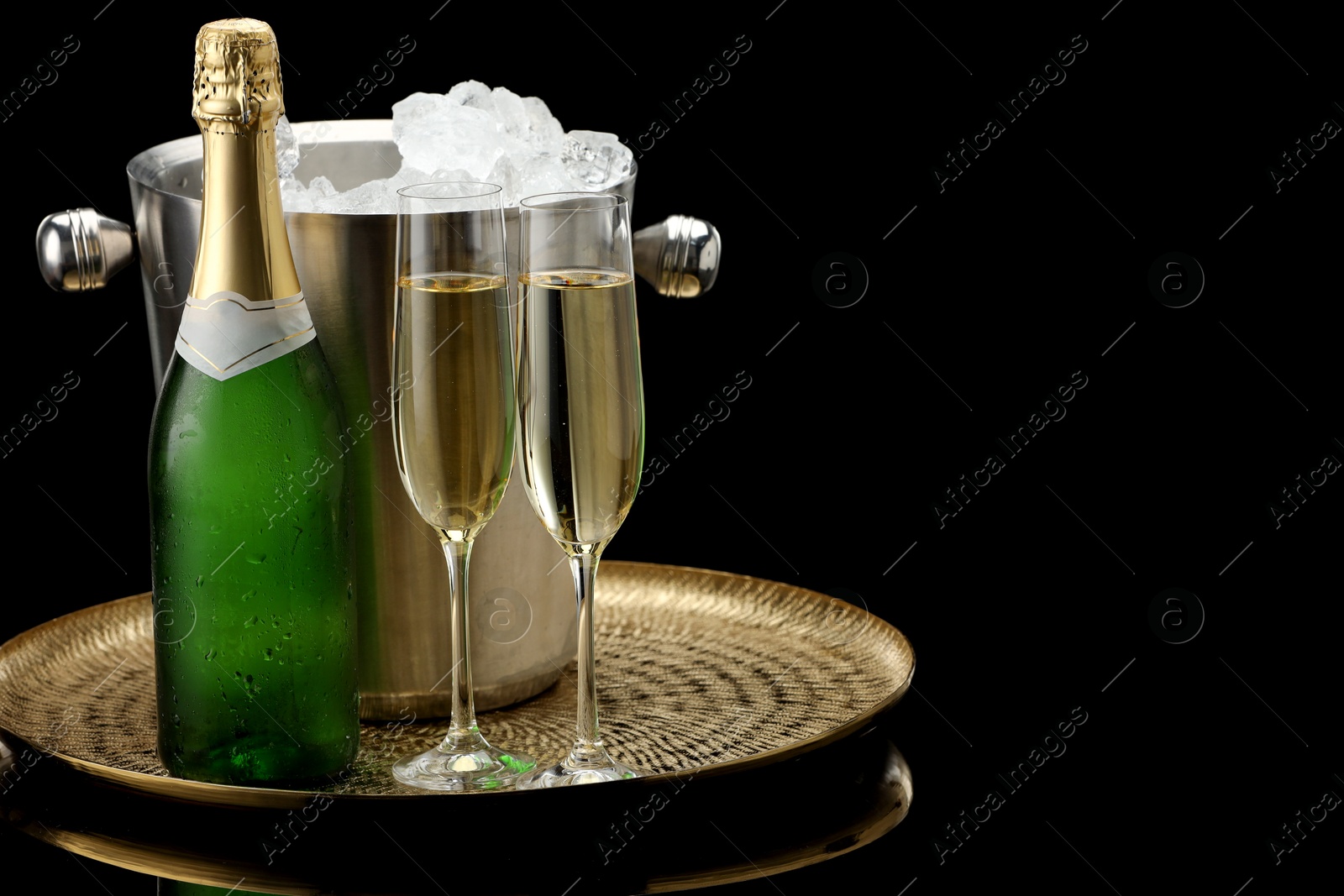 Photo of Bottle of sparkling wine, glasses and cooler bucket on black mirror surface. Space for text