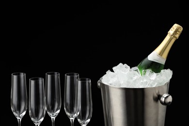 Photo of Bottle of sparkling wine in cooler bucket and glasses against black background, closeup. Space for text