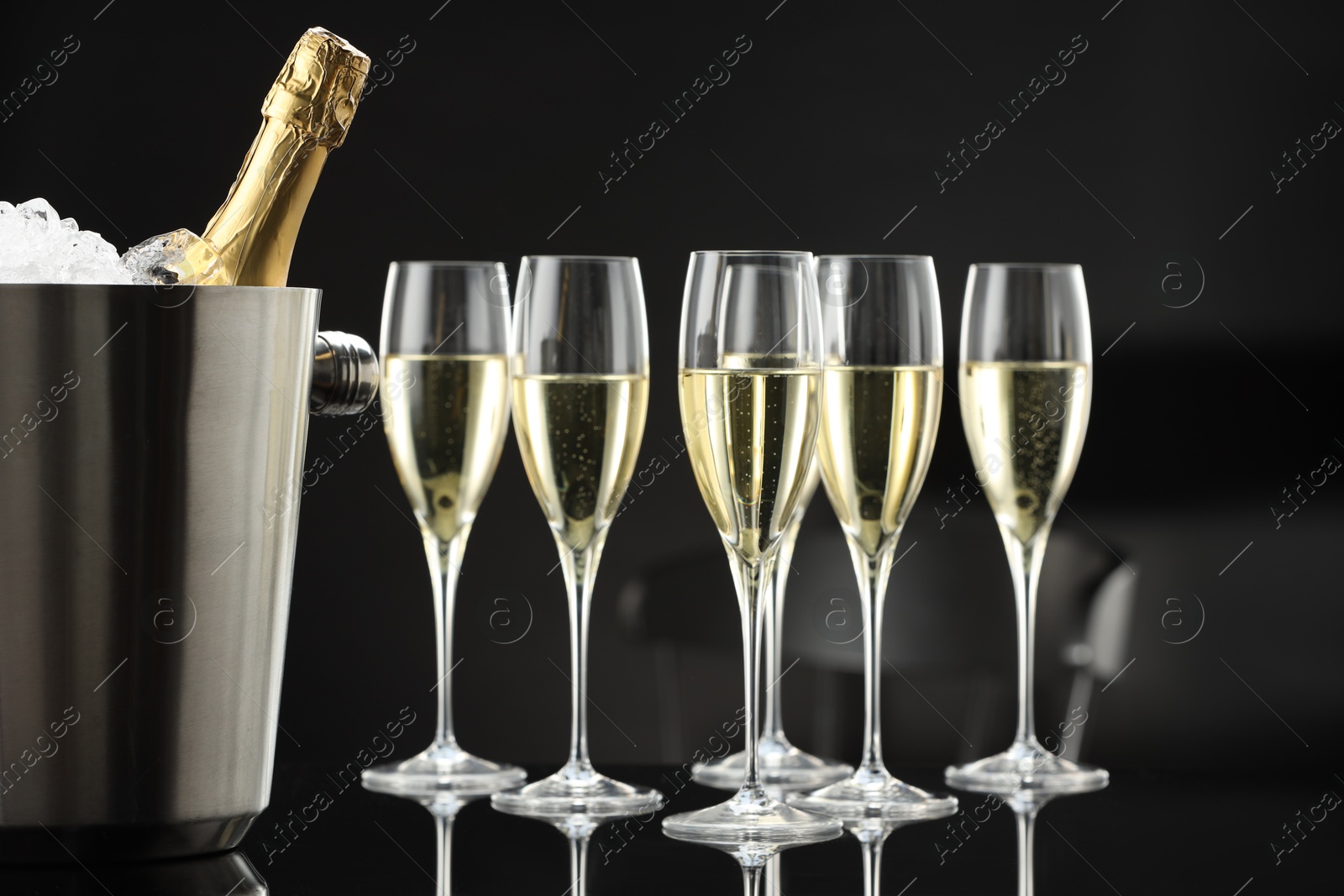 Photo of Bottle of sparkling wine in cooler bucket and glasses on black mirror surface