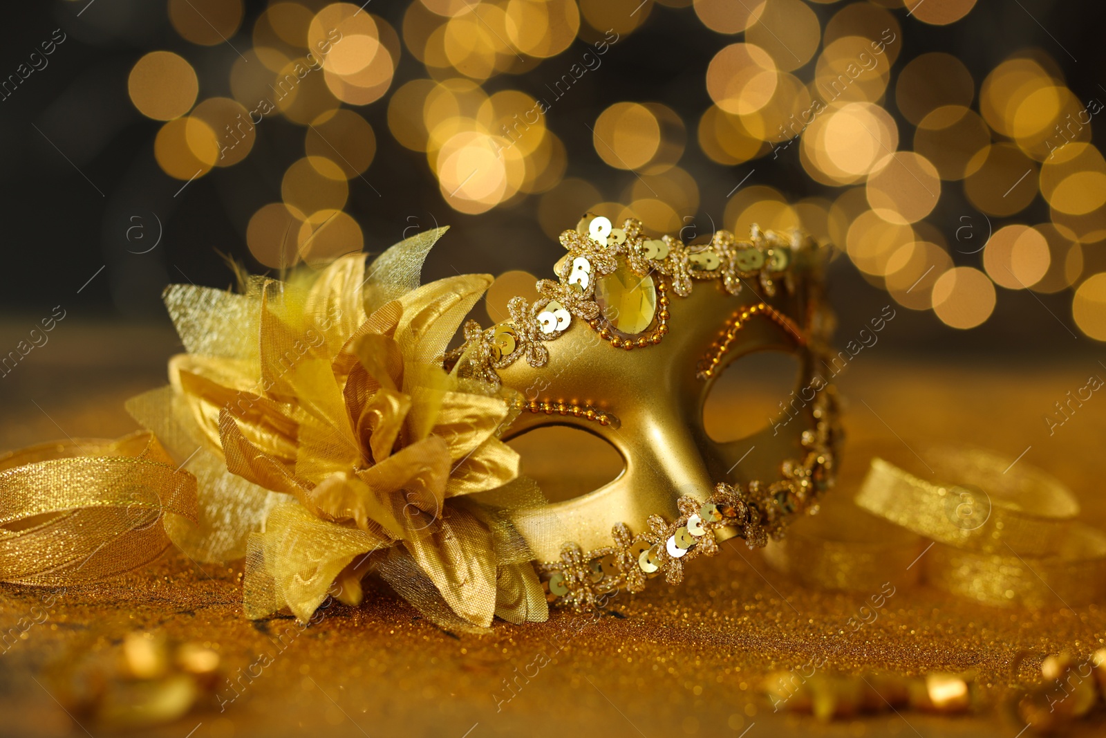 Photo of Beautiful carnival mask on golden shiny surface against blurred lights, closeup