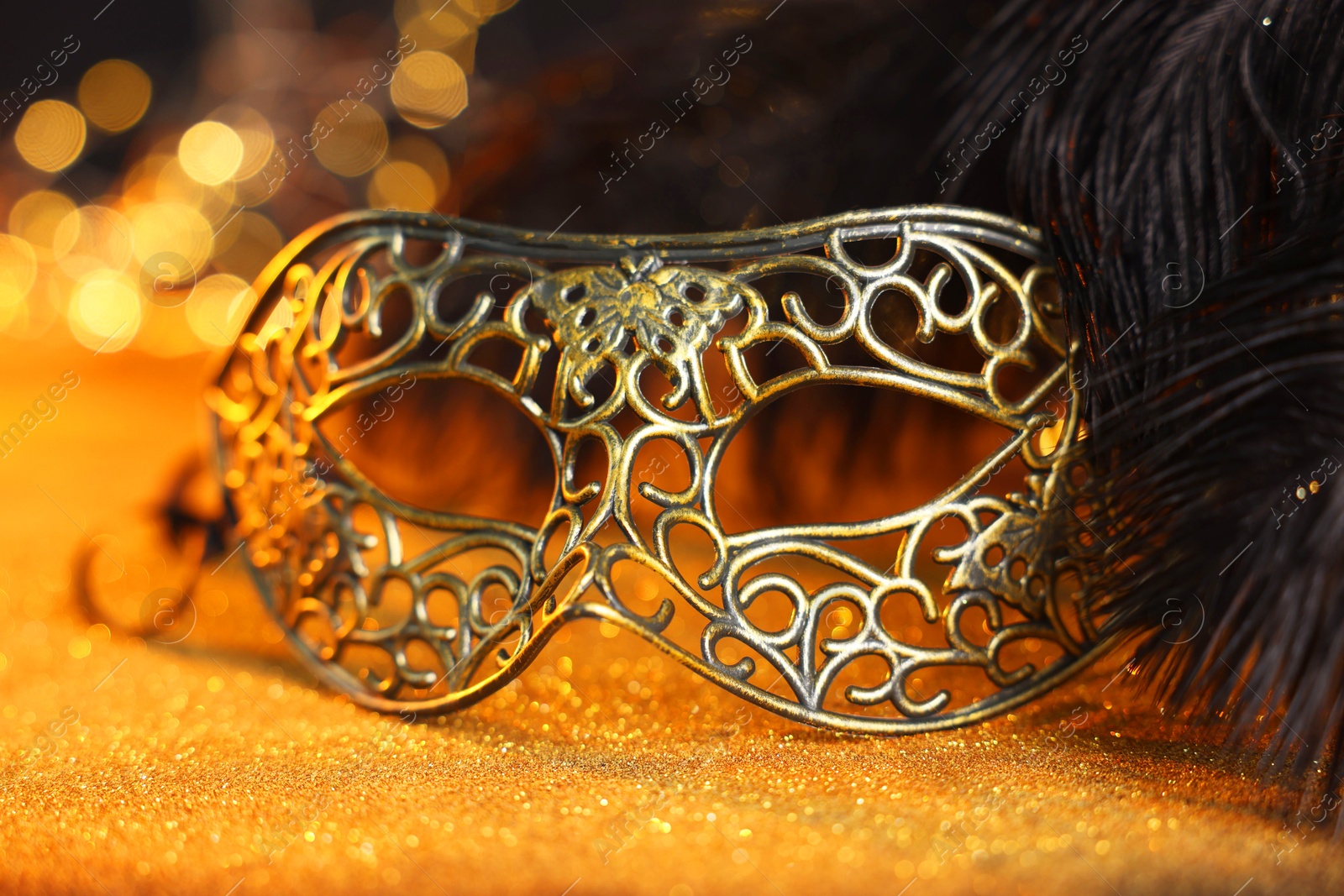 Photo of Beautiful carnival mask and black feathers on golden background against blurred lights, closeup. Bokeh effect