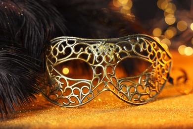 Photo of Beautiful carnival mask and black feathers on golden background against blurred lights, closeup. Bokeh effect