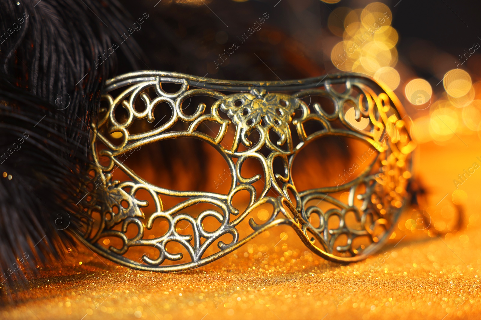 Photo of Beautiful carnival mask and black feathers on golden background against blurred lights, closeup. Bokeh effect