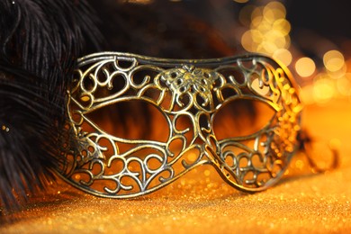 Photo of Beautiful carnival mask and black feathers on golden background against blurred lights, closeup. Bokeh effect