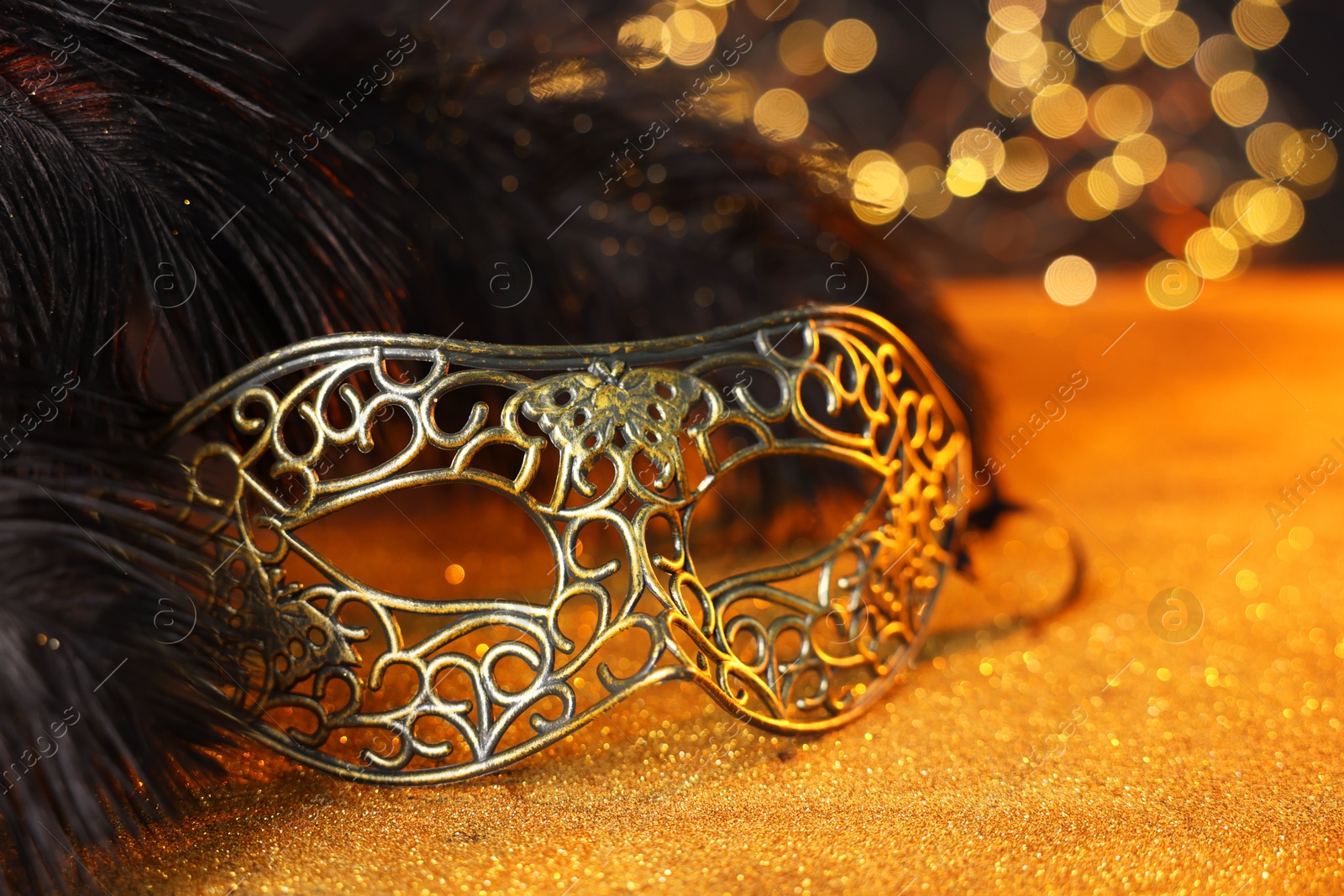 Photo of Beautiful carnival mask and black feathers on golden background against blurred lights, closeup. Bokeh effect