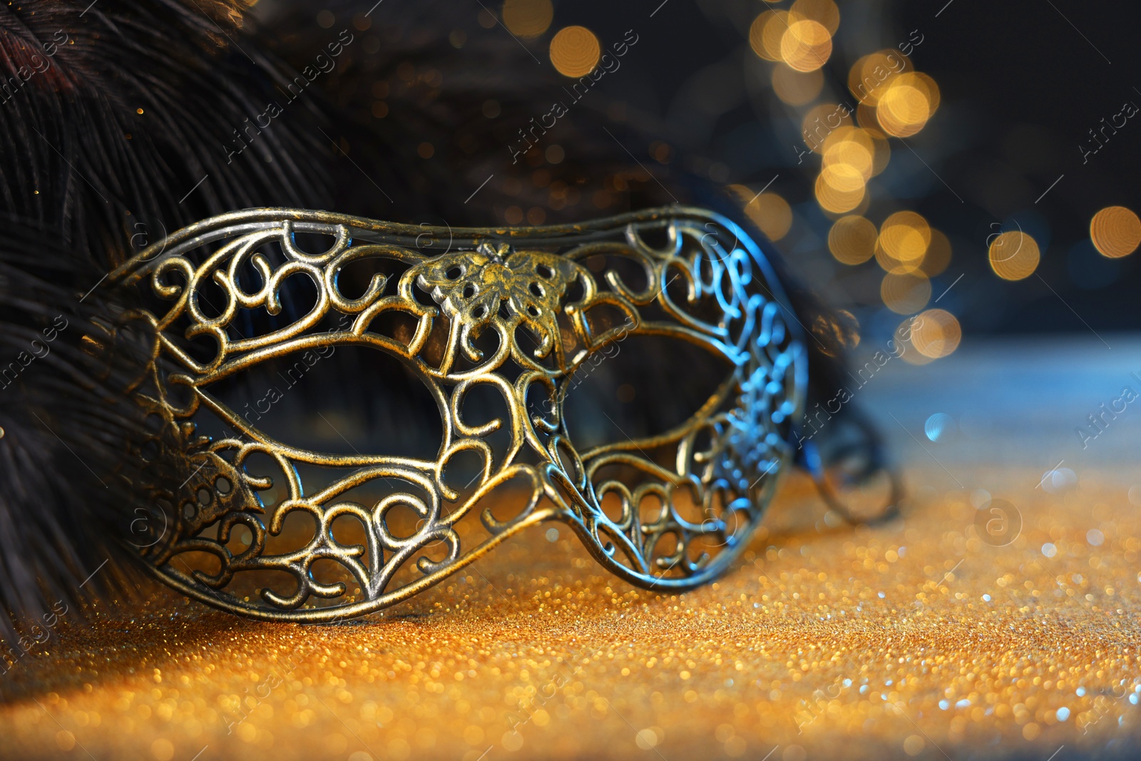 Photo of Beautiful carnival mask and black feathers on golden background against blurred lights, closeup. Bokeh effect