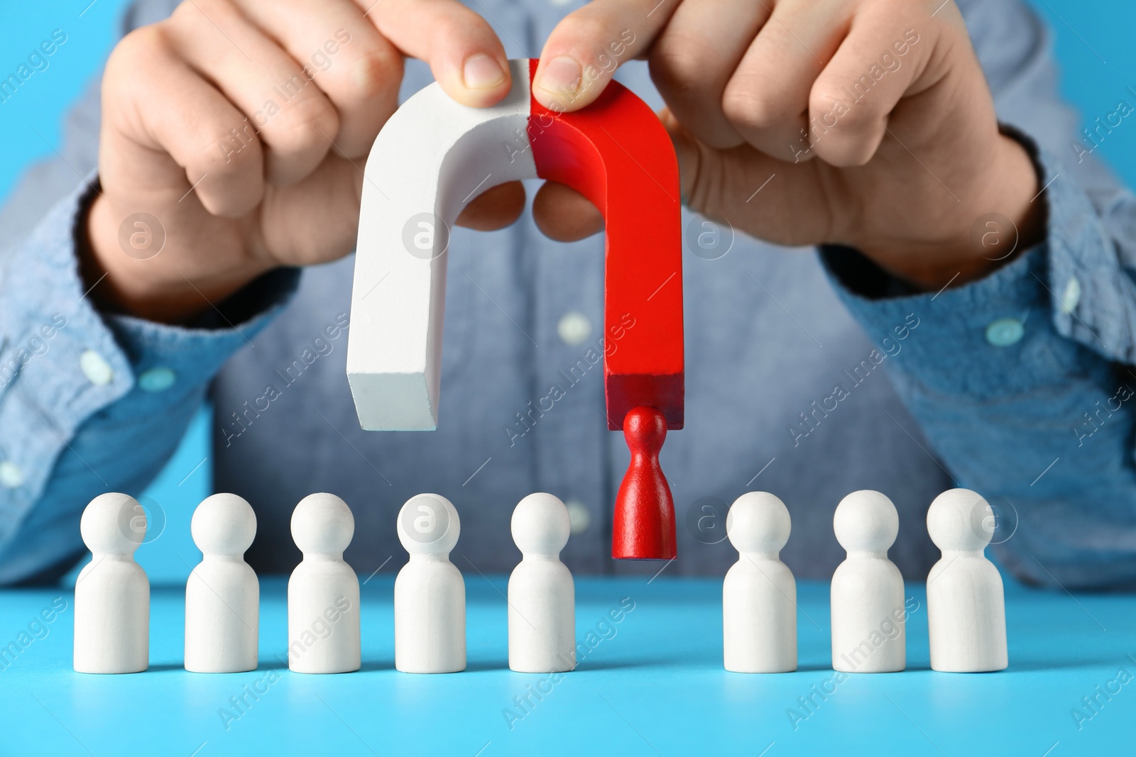 Photo of Man with magnet attracting red piece among wooden ones on light blue background, closeup