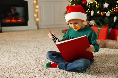 Photo of Cute little boy with Santa hat reading book on rug in room decorated for Christmas, space for text