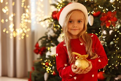 Photo of Little girl in Santa hat with Christmas ball at home, space for text