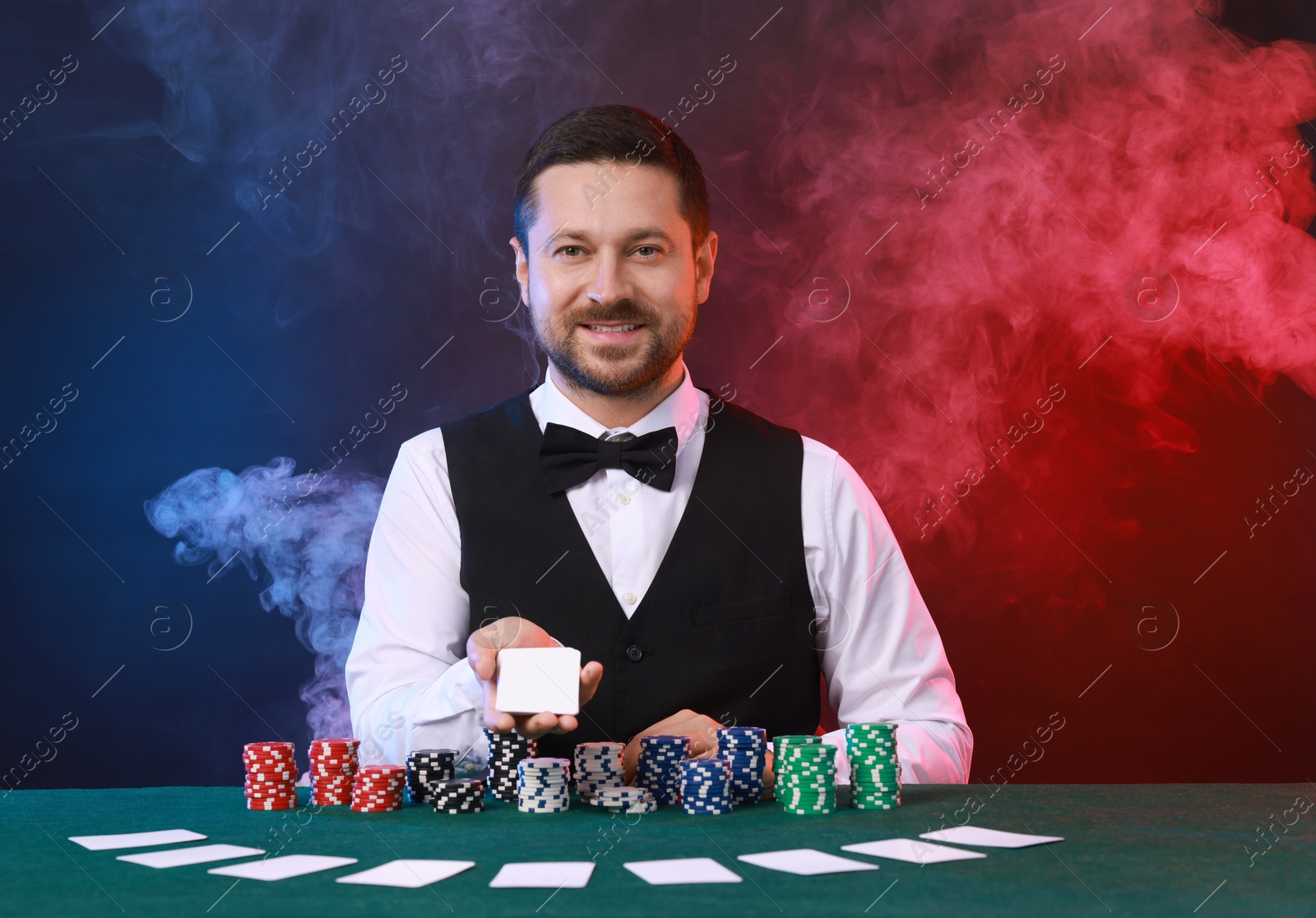 Photo of Professional croupier holding playing cards and casino chips at gambling table against color background with smoke