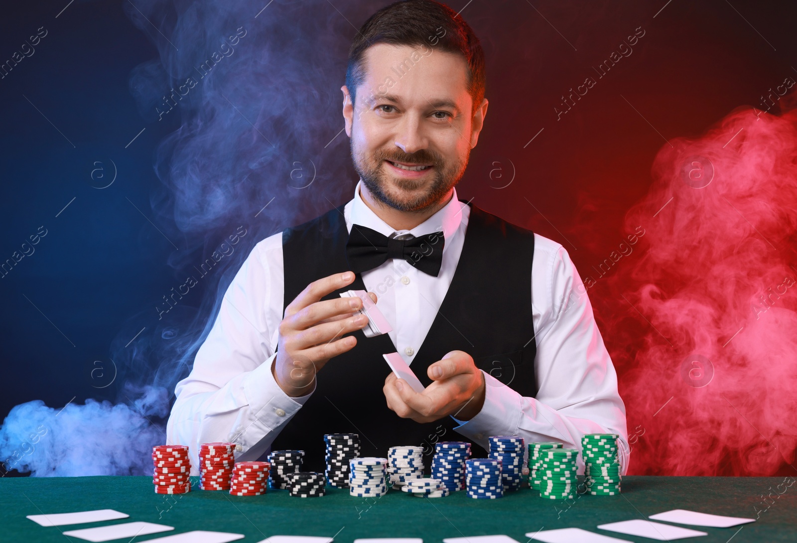 Photo of Professional croupier shuffling cards at gambling table against color background with smoke