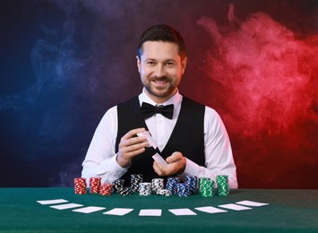 Professional croupier shuffling cards at gambling table against color background with smoke