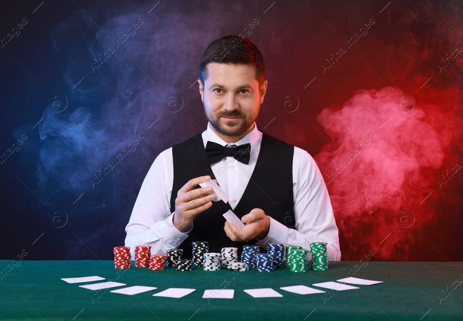 Photo of Professional croupier shuffling cards at gambling table against color background with smoke