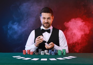 Professional croupier shuffling cards at gambling table against color background with smoke