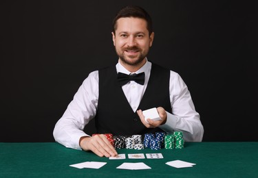 Professional croupier with playing cards at gambling table against black background